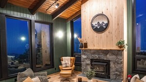 A cozy living room with a stone fireplace, wooden ceiling beams, and large windows offering a view of the outdoors. - Sunshine Ridge View - New Remodel - New Listing! (Anchorage)