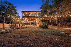A modern two-story building with a covered outdoor area, surrounded by trees and a grassy lawn. - Lake Travis Private Dock + Boat Slip + Hot Tub (Briarcliff)