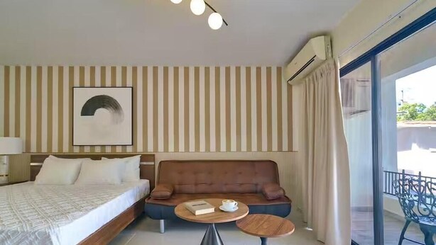 Une chambre d’hôtel avec un lit, un canapé, deux petites tables, une œuvre d’art accrochée au mur et une grande fenêtre avec des rideaux.