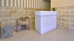 Un espace d’accueil avec un mur en pierre, un comptoir de réception blanc, deux chaises en bois et un lampadaire au motif décoratif.