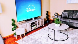 Een woonkamer met een flatscreen televisie waarop het Disney+-logo staat, een grijze bank, een ronde salontafel en een klein witte bijzettafeltje.