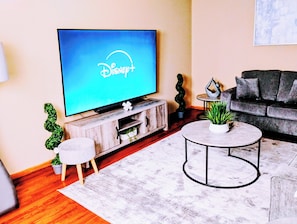 Una sala de estar con un televisor de pantalla plana mostrando el logotipo de Disney+, un sofá gris, una mesa de centro redonda y una mesita auxiliar blanca.