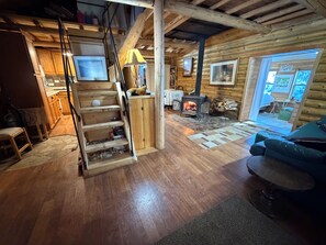 En stuga byggd av timmer med en interiör som inkluderar en trappa i trä, en öppen spis, en soffa och ett matbord.