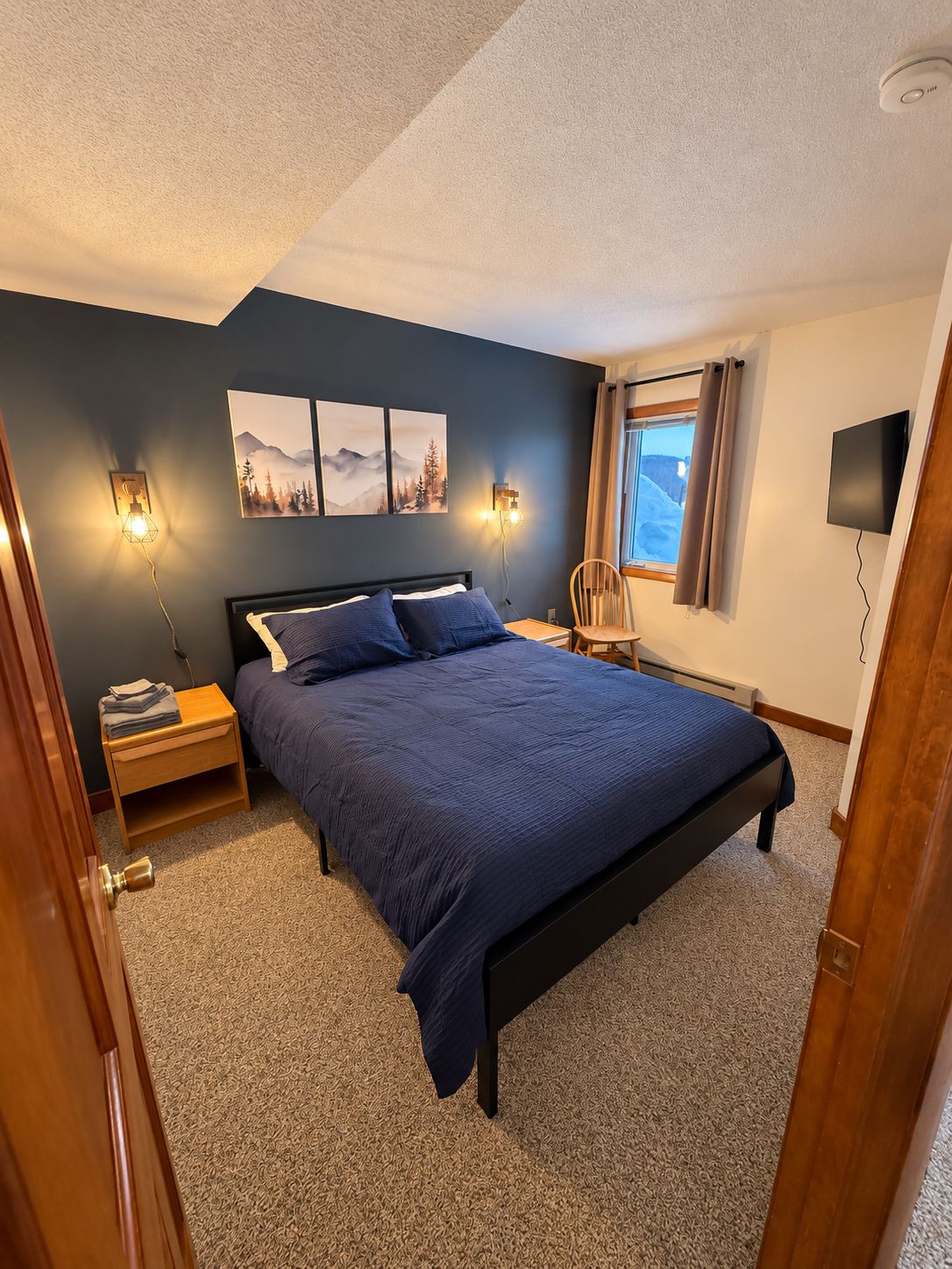 Ein Schlafzimmer mit einem Bett, Nachttischlampen, einem Nachttisch, einem Fenster mit Vorhängen und einem an der Wand befestigten Fernseher.