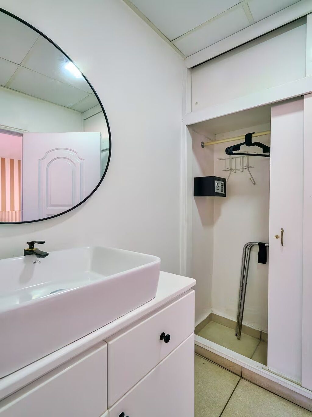 A bathroom with a white sink, a round mirror, a wardrobe with a door, and a shower area.