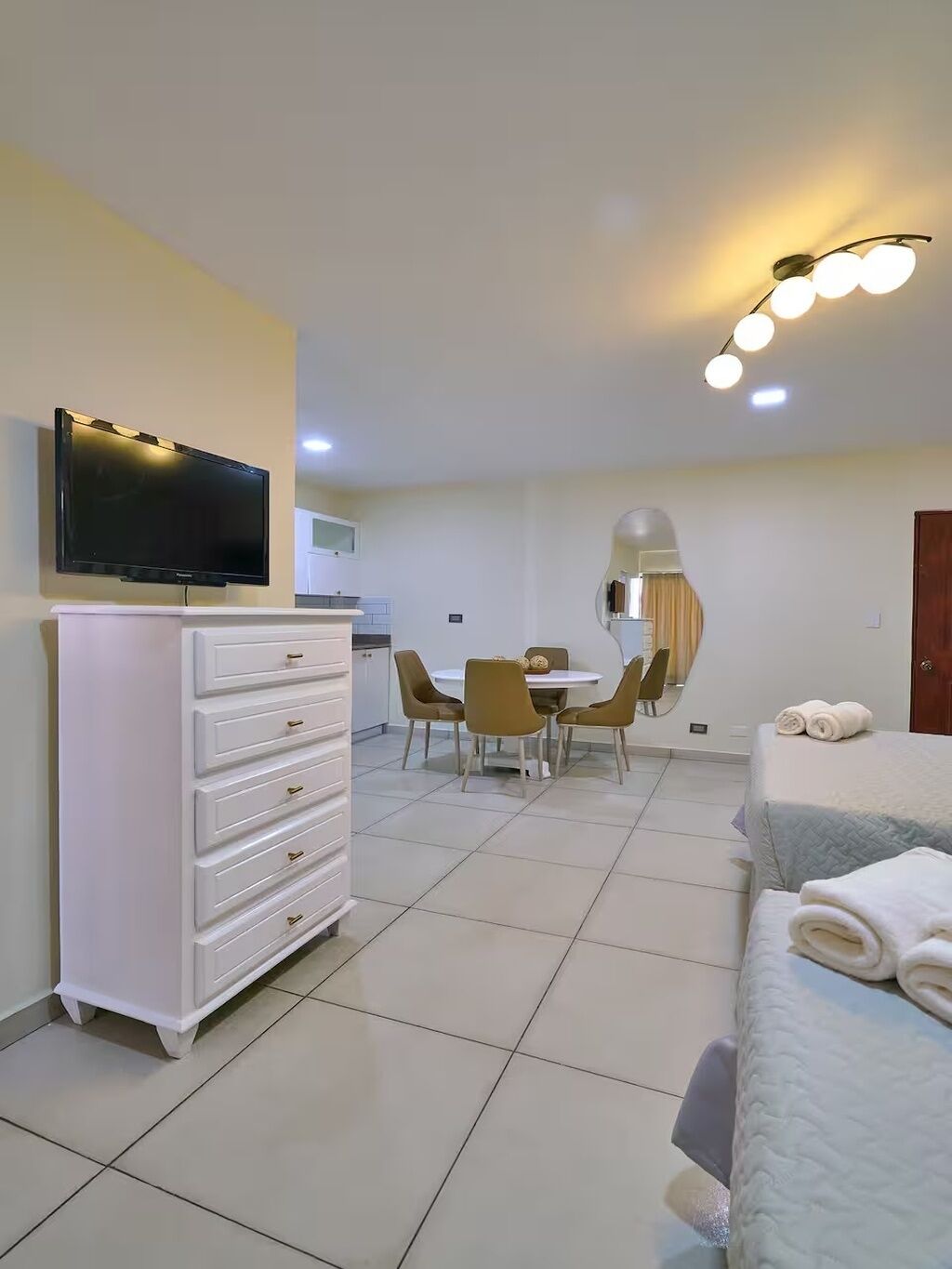 Habitación de hotel con televisor, cómoda, mesa de comedor con sillas, cama con toallas e iluminación en el techo.