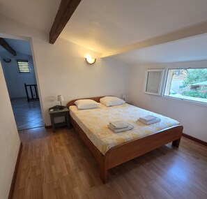 A bedroom with a wooden bed, a nightstand, a wall-mounted lamp, and a window with a view. - CASA FOCI -  maison T3 (Sainte Lucie de Porto Vecchio)