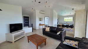 A modern living room with a flat-screen TV, a wooden coffee table, a black leather sofa, and a dining area with a kitchen in the background. - CASA FUNTA - villa T5 - vue mer- piscine (Sainte Lucie Porto-Vecchio)