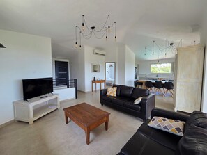 A modern living room with a flat-screen TV, a wooden coffee table, a black leather sofa, and a dining area with a kitchen in the background. - CASA FUNTA - villa T5 - vue mer- piscine (Sainte Lucie Porto-Vecchio)