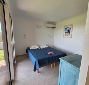 A single bed with a blue bedspread, a small table, an air conditioning unit, and a wall-mounted picture. - CASA FUNTA - villa T5 - vue mer- piscine (Sainte Lucie Porto-Vecchio)