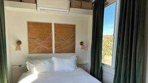 Een hotelkamer met een bed, twee wandlampen, een raam met gordijnen en een luchtverkoeler.