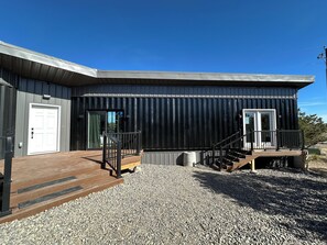 A modern building with a metal exterior, a wooden deck, and a gravel driveway. - Container Home at the Edge of the San Juans (South Fork)