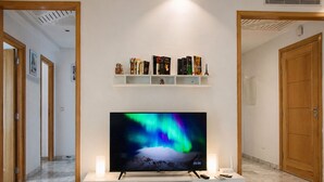 A TV on a white stand in a hallway with a shelf above it holding books and decorative items. - Luxury Oasis S 2 Comfort at the Garden in Carthage (Tunis)
