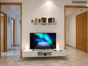 A TV on a white stand in a hallway with a shelf above it holding books and decorative items. - Luxury Oasis S 2 Comfort at the Garden in Carthage (Tunis)