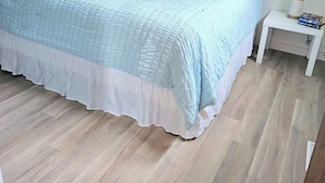 Una cama bien tendida con una colcha azul claro y detalles blancos, una mesita de noche blanca con una lámpara y libros, y suelo de madera.
