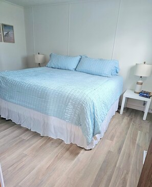 Una cama bien tendida con una colcha azul claro y detalles blancos, una mesita de noche blanca con una lámpara y libros, y suelo de madera.