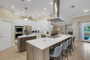 A modern kitchen with white cabinets, stainless steel appliances, a central island with bar stools, and a ceiling fan. - Elegant 4-bed Home w/ Pool & Spa Near Anna Maria (West Bradenton)