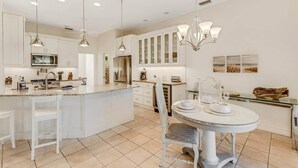A modern kitchen with white cabinetry, a central island, pendant lighting, and a dining area with a round table and chairs. - Elegant 4-bed Home w/ Pool & Spa Near Siesta Key (Bee Ridge)