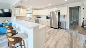A modern kitchen with white cabinetry, a stainless steel refrigerator, and a bar area with stools. - Huge Home- 1 min Walk to Beach- Perfect Location (Surf City)
