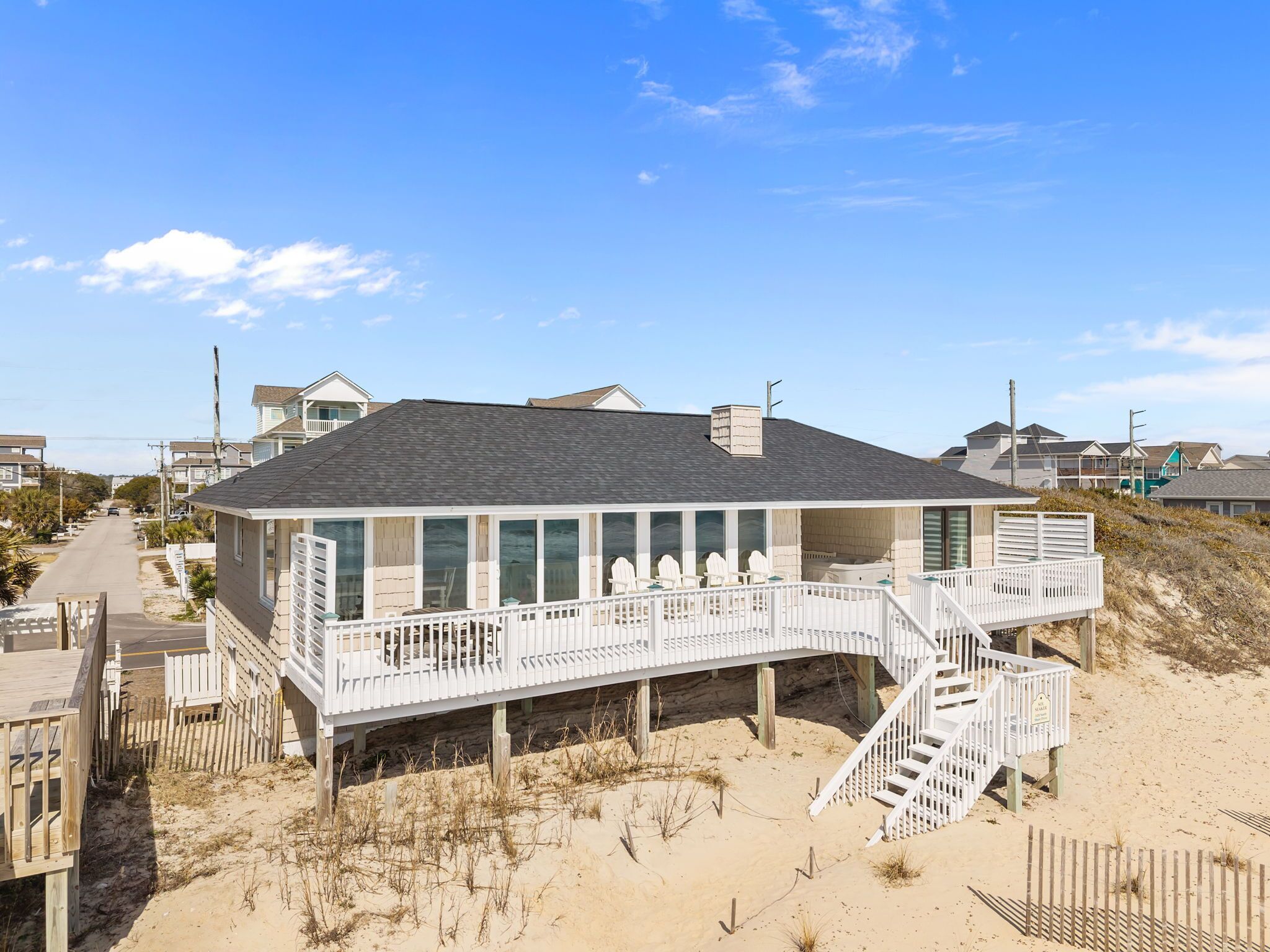 A beachfront house with a covered deck, a staircase leading to a balcony, and a sandy beach in the background.