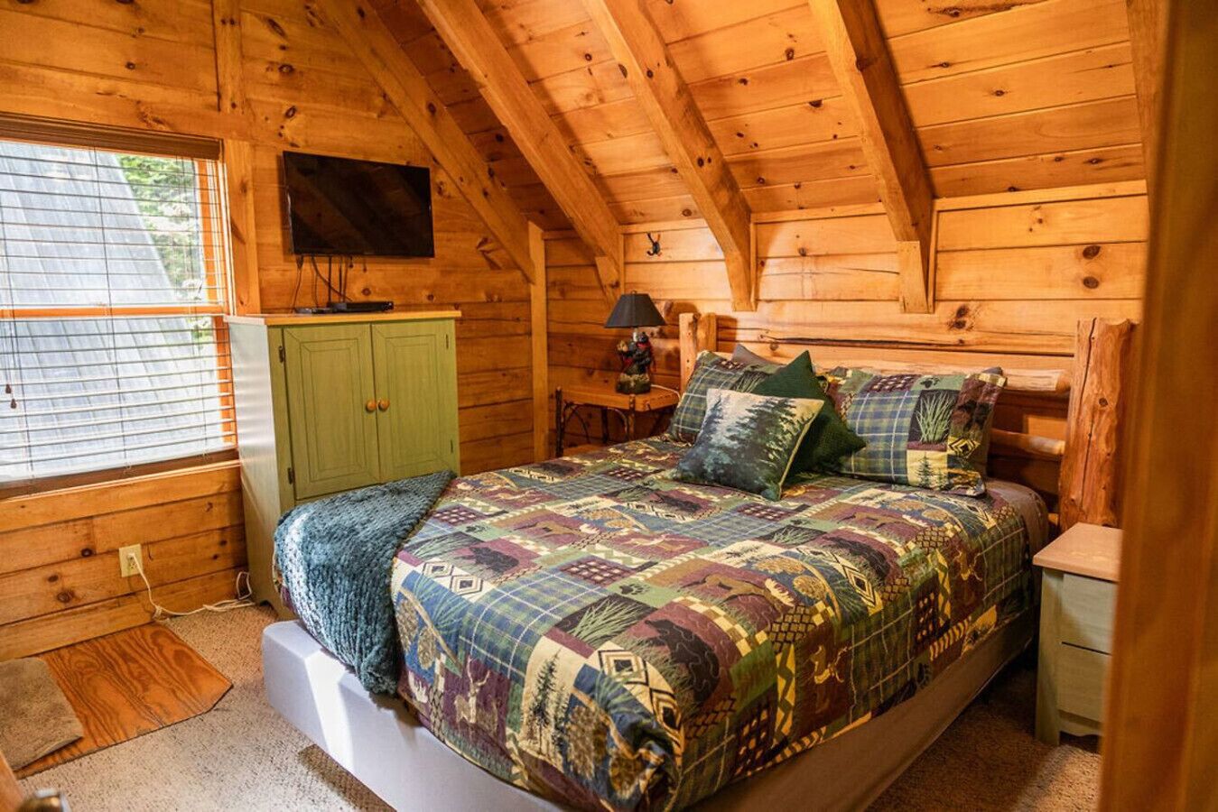 Ein Schlafzimmer einer Holzhütte mit einem Bett, einem grünen Schrank, einem Nachttisch mit Lampe und einem an der Wand befestigten Fernseher.
