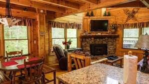 A log cabin interior with a stone fireplace, a television mounted above it, and a dining area with wooden furniture. - Rustic Pet Friendly Cabin in Pigeon Forge! (Pigeon Forge)
