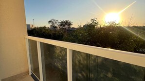 Un balcón con barandilla de vidrio, cielo despejado y el sol poniéndose al fondo.