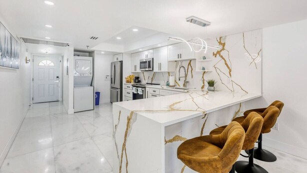 A modern kitchen with white cabinetry, a marble island, and a bar area with velvet chairs. - Oceanview Oasis w/ Balcony | Steps to the Sand (Madeira Beach)