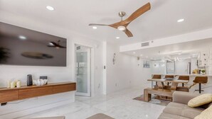 A modern living room with a flat-screen TV, a ceiling fan, a dining area with a table and chairs, and a sofa. - Oceanview Oasis w/ Balcony | Steps to the Sand (Madeira Beach)