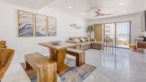 A modern living room with a wooden dining table, a beige sofa, and a large window offering a view. - Oceanview Oasis w/ Balcony | Steps to the Sand (Madeira Beach)