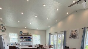 A spacious living room with a large sectional sofa, a ceiling fan, recessed lighting, a kitchen area, and a dining table.