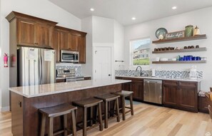 Una cocina moderna con armarios de madera, electrodomésticos de acero inoxidable, una isla de mármol con taburetes y estantes decorativos.