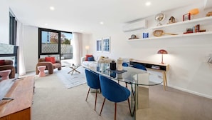 A modern living space with a glass dining table, blue chairs, a sofa, and a bookshelf.