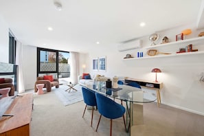 A modern living space with a glass dining table, blue chairs, a sofa, and a bookshelf. - Stylish 1-Bed Retreat in Braddon (Braddon)