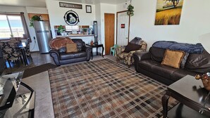 A living room with a sofa, a coffee table, a patterned rug, and a kitchen area with a refrigerator and a window.