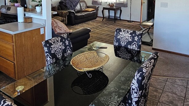 A dining area with a glass table and patterned chairs, a kitchen with wooden cabinets, a ceiling fan, and a living room with a sofa and a window.