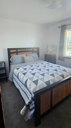 A bedroom with a bed, a nightstand, a bedside table, a window with curtains, and a ceiling fan.