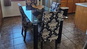 A dining area with a glass table, patterned chairs, a chandelier, a refrigerator, and wooden cabinets.