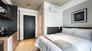 Una habitación de hotel moderna con una cama, una kitchenette pequeña, una puerta y un equipo de aire acondicionado.