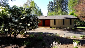 A small, single-story building with a red door and beige siding, surrounded by trees and shrubs. - Family Seattle Stay • Swim Spa • Fire Pit • 9 beds (Woodinville)