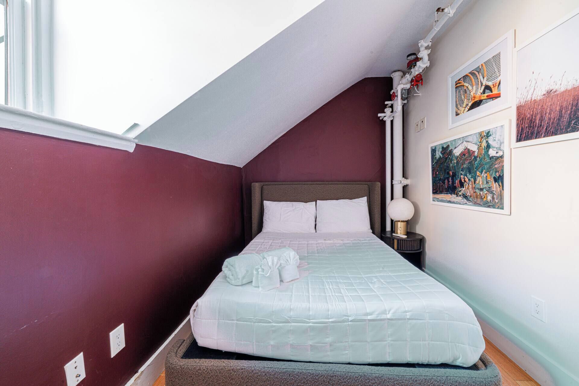Ein Schlafzimmer mit schrägen Wänden, einem Bett mit weißer Bettwäsche, einem Nachttisch mit einer Lampe und zwei gerahmten Bildern an der Wand.
