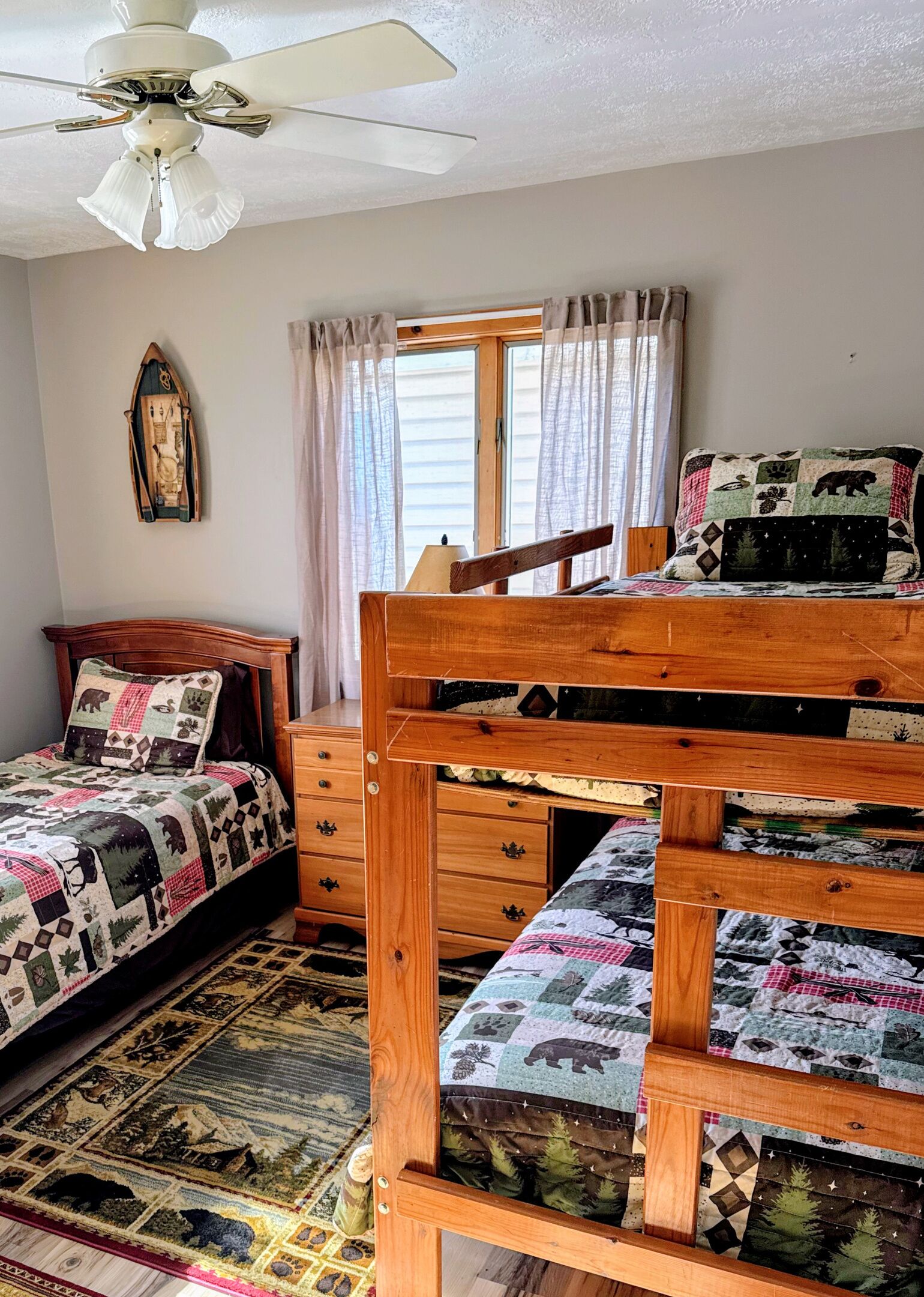 Un lit superposé avec une tête de lit matelassée, une commode en bois avec des tiroirs, un ventilateur de plafond, une fenêtre avec des rideaux et un élément de décoration fixé au mur.
