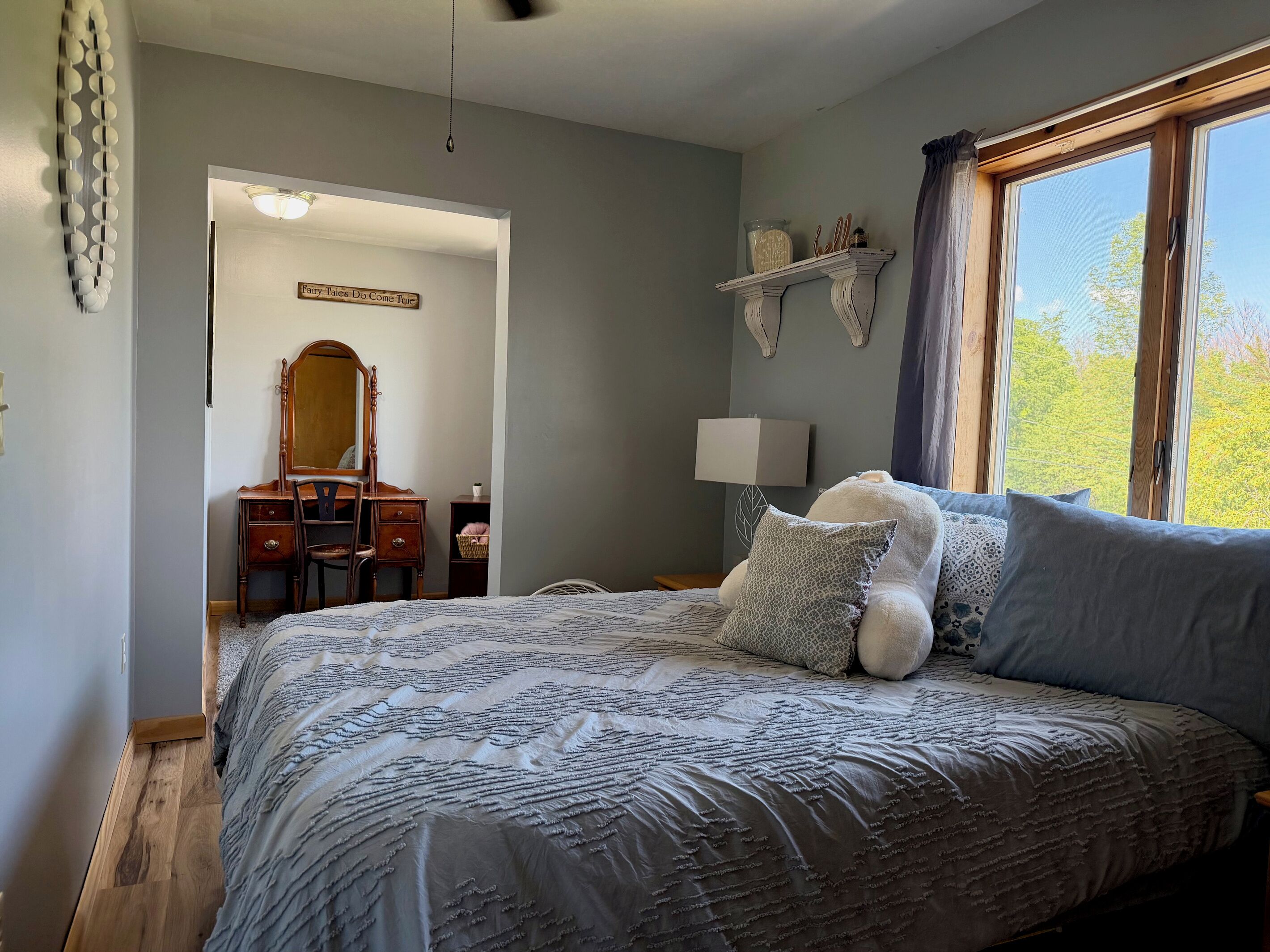 A bedroom with a large bed, a dresser, a mirror, a lamp, and a window with a view of trees.