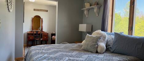 A bedroom with a large bed, a dresser, a mirror, a lamp, and a window with a view of trees.