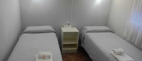 Una habitación de hotel pequeña y sencilla con dos camas individuales, una mesita de noche, aire acondicionado de pared y una ventana con cortinas.