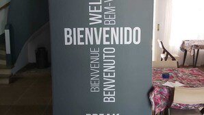 Uma placa de boas-vindas com 'WELCOME' e 'BIENVENIDO' em vários idiomas, uma escadaria e uma área de refeições com mesa e cadeiras.