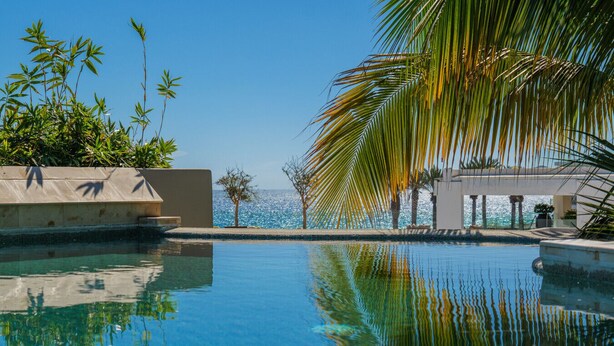 Ein klares, blaues Schwimmbecken mit Spiegelungen, Palmen und Blick aufs Meer.
