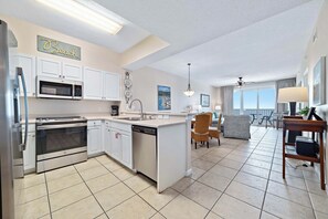 A modern kitchen with stainless steel appliances, a central island, and a dining area with wicker furniture. - Windemere 1103 | Beachfront | Seasonally Heated Pool | My Beach Getaways (Pensacola)