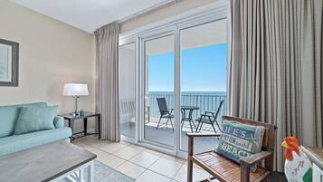 A hotel room with a sofa, a table, a lamp, a wooden chair with a cushion, and a view of the sea through a sliding glass door.