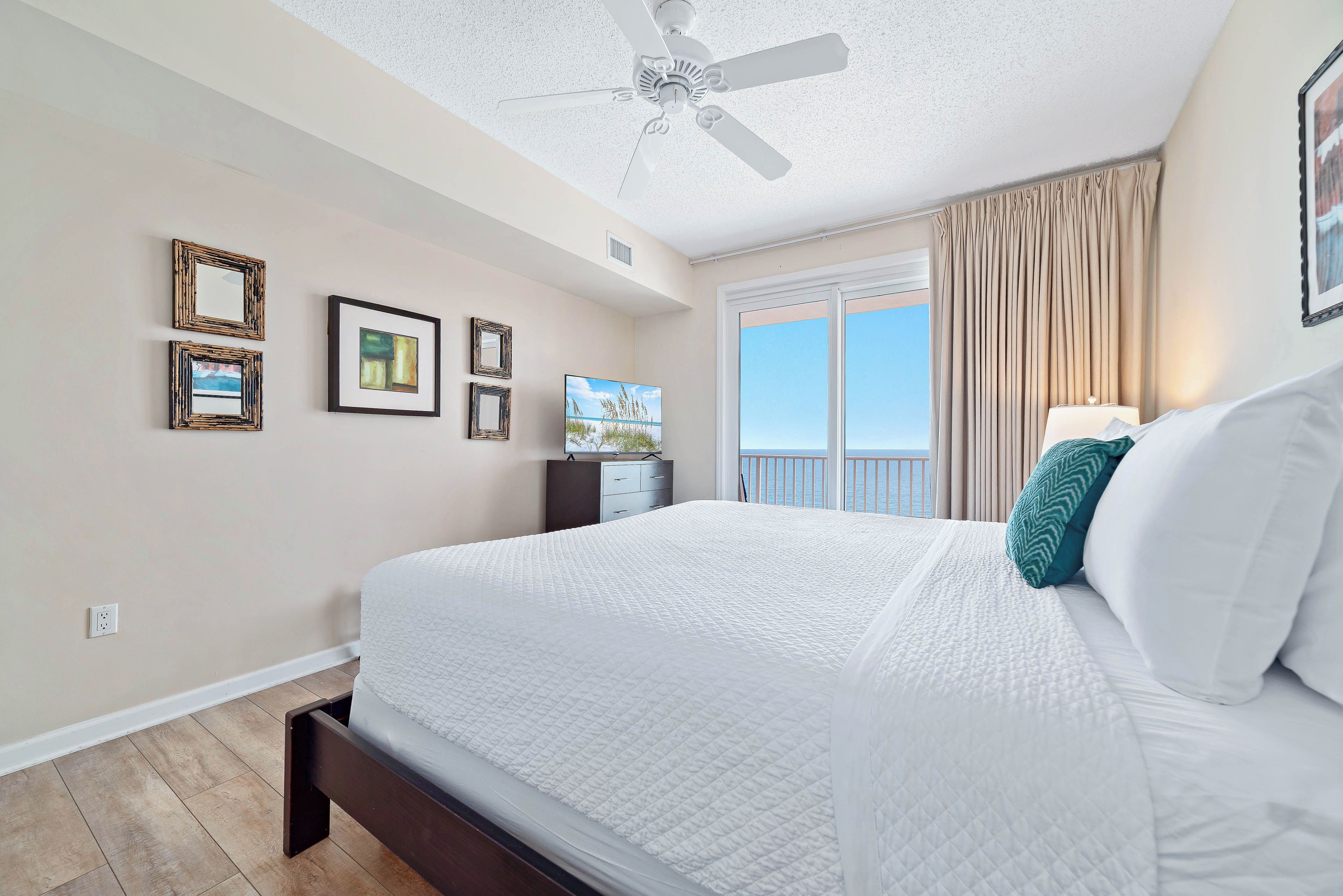 A hotel room with a bed, ceiling fan, artwork on the wall, and a window with curtains.